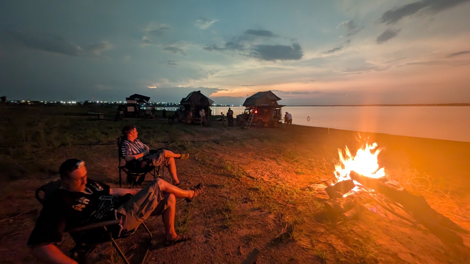 Overlanders relaxing around a campfire beside the river at night